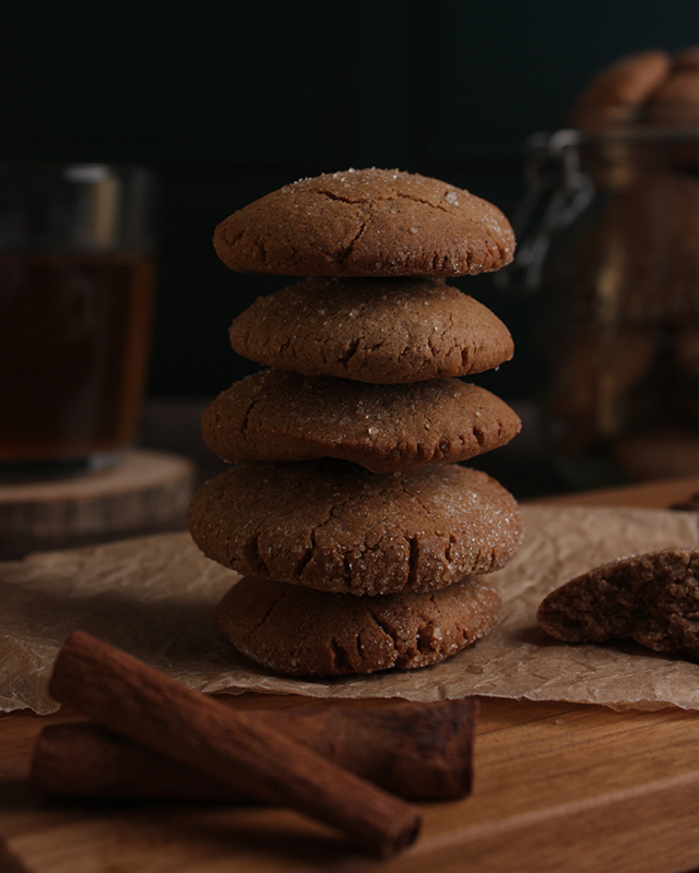 Une pile de biscuit à la cannelle sur une planche en bois. En arrière plan un thé et un bocaux de biscuits. Au premier plan deux bâtons de cannelle